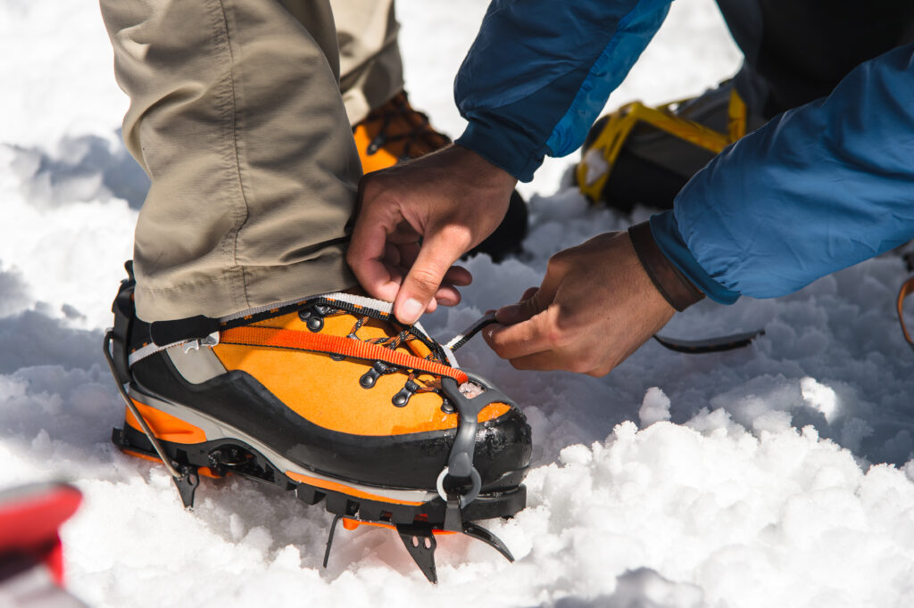 alpiniste en train d'installer une paire de crampon sur des chaussures de haute montagne. Il est possible de faire une réparation sur ses chaussures de randonnée